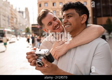 Un joyeux couple gay multiethnique partage un moment de lumière, avec un partenaire tenant un appareil photo vintage, alors qu'ils embrassent dans un cadre urbain animé. Banque D'Images