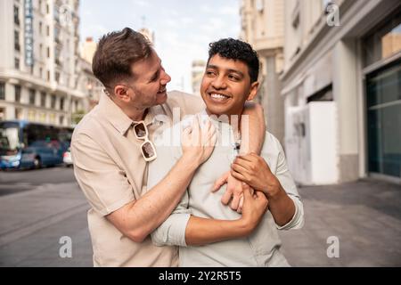 Un joyeux couple gay multiethnique partage une étreinte chaleureuse et sourit dans une rue de la ville, leur bonheur évident alors qu'ils chérissent leur temps ensemble. Banque D'Images