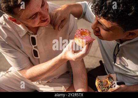 Une scène touchante d’un couple gay multiethnique partageant intimement des beignets par une journée ensoleillée, soulignant l’affection et la connexion entre eux. Banque D'Images