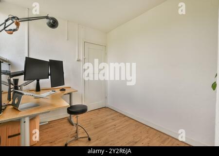 Un bureau à domicile minimaliste lumineux avec un bureau en bois, deux moniteurs et un tabouret roulant. L'espace est bien rangé avec des murs blancs et un floori en bois naturel Banque D'Images