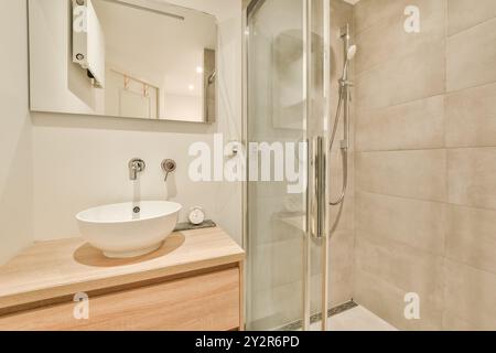 Cette image présente une salle de bains moderne avec une cabine de douche en verre, un lavabo blanc élégant sur une vanité en bois, avec des carreaux de couleur neutre. Banque D'Images