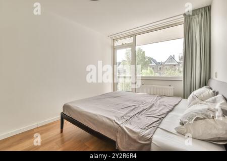 Une chambre confortable et minimaliste avec un lit double avec literie grise, planchers de bois franc, et une grande fenêtre avec des rideaux fluides laissant entrer ample daylig Banque D'Images