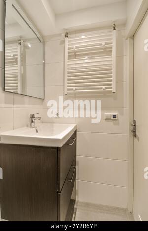 Une salle de bains contemporaine avec un lavabo en céramique blanche, un meuble-lavabo en bois sombre et des stores vénitiens sur la fenêtre. Banque D'Images