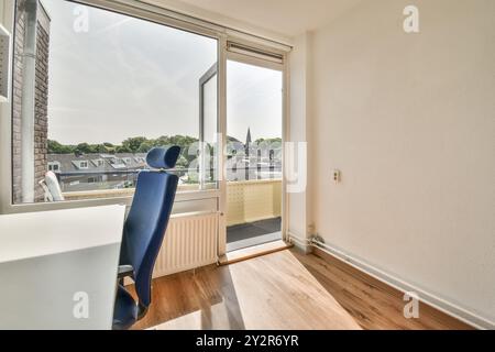 Une pièce inoccupée avec un sol stratifié, une fenêtre ouverte et une chaise de bureau bleue unique. La lumière du jour remplit l'espace, offrant une vue sur les toits. Banque D'Images