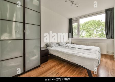 Une chambre moderne confortable avec un lit double avec literie grise, grande armoire en verre dépoli, parquet et une fenêtre avec vue sur la verdure. Banque D'Images