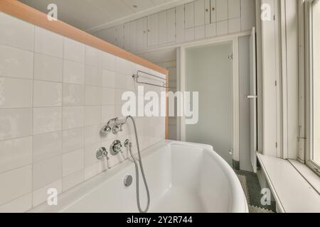 La salle de bains élégante et contemporaine est dotée d'une baignoire en céramique blanche avec robinets argentés, murs carrelés blancs et touches de bois. Banque D'Images