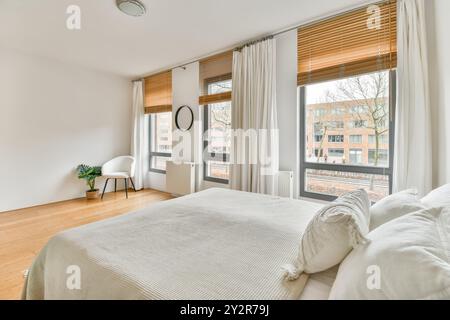 Cette chambre moderne lumineuse et aérée comprend de grandes fenêtres avec stores et un lit confortable. Banque D'Images