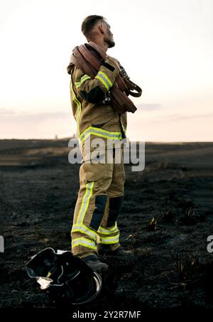 Un pompier se tient debout dans une forêt carbonisée, tenant un gros tuyau d'incendie sur son épaule au crépuscule, représentant la bravoure et les suites de Banque D'Images