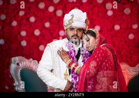 Un couple vêtu d'une tenue de mariage indienne traditionnelle posant sur un fond floral rouge Banque D'Images