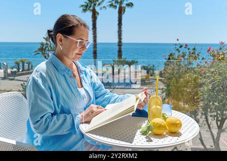 Femme mature satisfaite lisant un livre sur le balcon de l'hôtel en été et contemplant la mer. Concept de détente et de vacances d'été dans le méditerr Banque D'Images