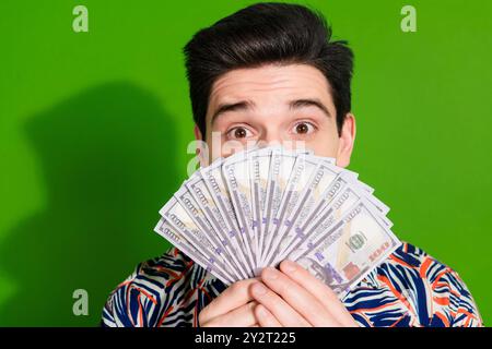 Photo de gars impressionné étonné avec chemise d'impression habillée de coupe de cheveux élégante cachant le visage derrière des dollars isolé sur fond de couleur verte Banque D'Images