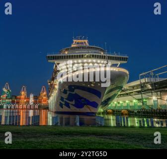 Un grand navire de croisière accostait dans un port la nuit avec des lumières brillantes se reflétant sur l'eau et les conteneurs de fret en arrière-plan. Banque D'Images