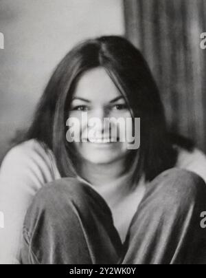 Photo en noir et blanc d'une jeune femme aux cheveux longs, souriante et assise avec les jambes tirées près de sa poitrine vers la fin des années 1960 ou le début des années 1970 Banque D'Images