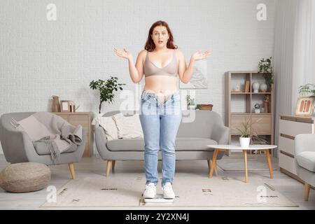 Surpoids jeune femme choquée en jeans serrés avec des balances à la maison. Concept de gain de poids Banque D'Images