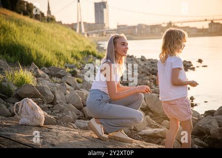 Par une chaude soirée d’été à Osijek, en Croatie, deux enfants profitent du bord de la rivière tandis que l’un d’eux s’agenouille au bord de l’eau. Le coucher de soleil doré baigne la scène, créa Banque D'Images