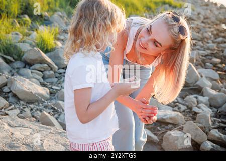 Lors d’une agréable soirée d’été à Osijek, en Croatie, une femme partage un moment léger avec un enfant au bord de la rivière, entourée de pierres et de verdure Banque D'Images