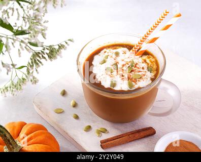 Latte végétalien à la citrouille épicée surmonté de crème fouettée à base de plantes et de graines de citrouille dans une tasse en verre sur une planche à découper blanche avec des pailles à boire Banque D'Images