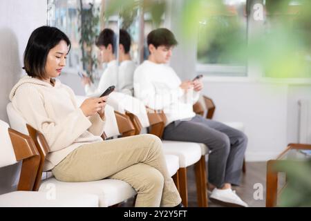 La fille est assise dans le hall du bureau de l'emploi, attendant l'entrevue ou la conversation avec le consultant de carrière. Regarde les actualités sur Internet via smartphone Banque D'Images