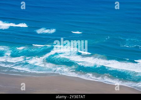 Une vue aérienne des vagues de l'océan avec des calottes blanches soufflées par le vent roulant sur une plage de sable. Banque D'Images