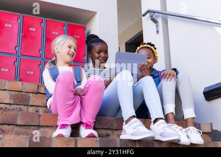 À l'école, assis dans les escaliers, trois filles multiraciales utilisant la tablette et souriant ensemble Banque D'Images