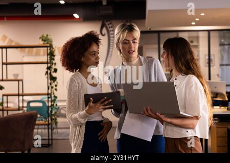 Collaborant dans le bureau, trois femmes multiraciales discutant du projet à l'aide d'un ordinateur portable et d'une tablette Banque D'Images