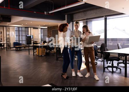 Collaborant dans un bureau moderne, trois femmes multiraciales discutent du travail à l'aide d'ordinateurs portables et de tablettes Banque D'Images