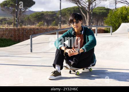 Assis sur une planche à roulettes, adolescent asiatique se relaxant au skatepark, profitant du temps libre en plein air Banque D'Images