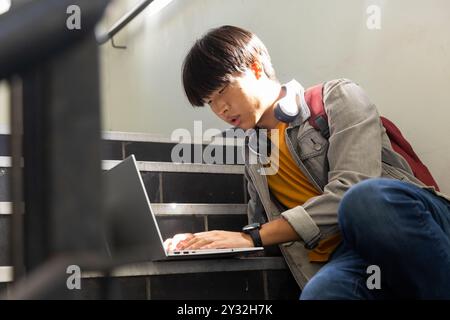 À l'école, adolescent asiatique assis dans les escaliers à l'aide d'un ordinateur portable avec des écouteurs autour du cou Banque D'Images