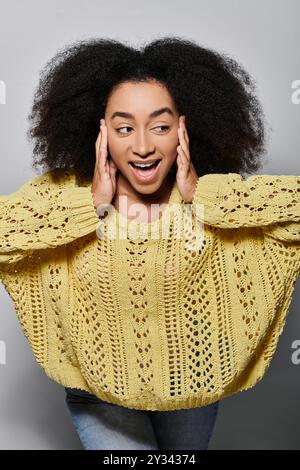 Une jeune femme vibrante aux cheveux bouclés sourit largement, mettant en valeur l'excitation dans son élégant pull jaune. Banque D'Images