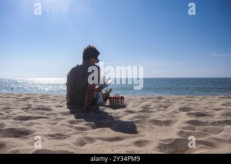 Jeune homme lit un livre sur la plage assis devant la mer bleue au coucher du soleil portant un T-shirt et des lunettes de soleil avec un panier de fruits à côté de lui. Banque D'Images