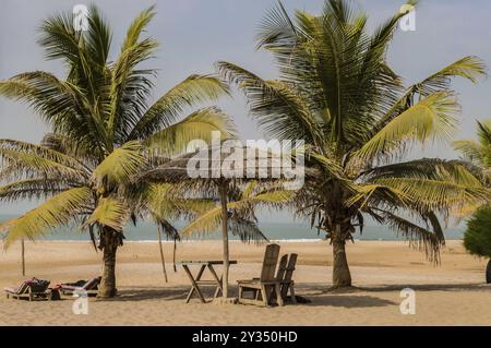 Deux chaises et une table basse sous une feuille de palmier et un parasol en bois sur la plage de Bijilo en Gambie Banque D'Images