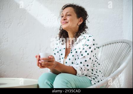 Une femme sereine savourant du café, assise dans une chaise confortable. Moment lumineux et paisible de détente capturé dans un cadre ensoleillé. Banque D'Images