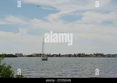 Large vue sur l'eau vers la baie avec un voilier blanc avec des arbres et des bâtiments au loin. Près du rivage avec de petites vagues vers la beac Banque D'Images
