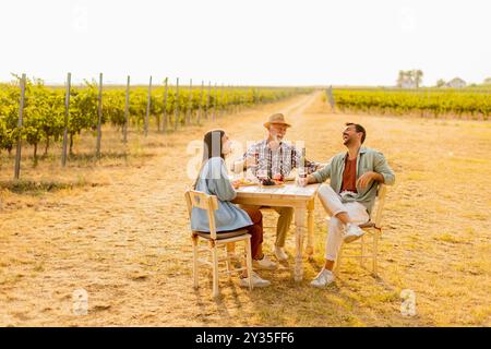 Trois amis se réunissent autour d'une table rustique dans un vignoble, dégustant du vin, des fruits frais, et la compagnie de l'autre sur une journée ensoleillée remplie de joie et de lau Banque D'Images
