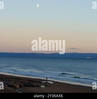 Un homme marche seul le long de la plage à Ogmore by Sea, Vale of Glamorgan, au coucher du soleil à la fin de l'été. Demi-lune. Banque D'Images