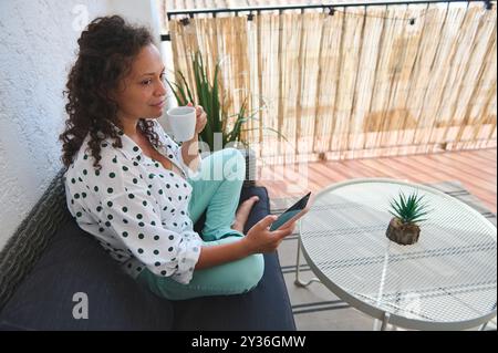 Une femme se détend sur un patio extérieur, en sirotant du café et en utilisant une tablette. Le cadre est serein et ensoleillé, mettant l'accent sur les loisirs et l'utilisation de la technologie. Banque D'Images