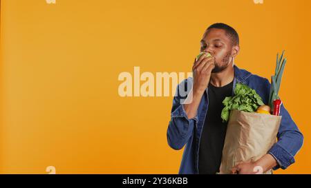 Mec végétalien afro-américain prenant une bouchée d'une pomme verte naturelle mûre, mangeant des fruits biologiques fraîchement récoltés en studio. Personne faisant la publicité de l'agriculture locale et des produits d'origine éthique. Caméra B. Banque D'Images