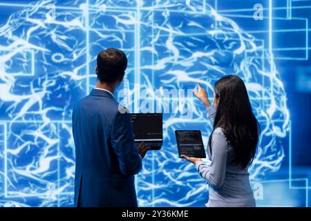 Équipe d'administrateurs utilisant tablette et ordinateur portable pour effectuer la maintenance sur les réseaux neuronaux d'intelligence artificielle. Membres du personnel INFORMATIQUE travaillant en équipe utilisant le système d'IA pour aider au traitement et à la transmission de l'information Banque D'Images