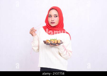 Femme asiatique portant un élégant hijab regarde vers la gauche, ramassant avec des baguettes et tenant une assiette contenant des sushis (nourriture japonaise). Beau Mus Banque D'Images