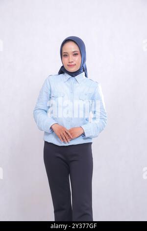 Jeune femme de bureau asiatique portant une chemise bleue hijab debout souriante élégamment les mains devant le nombril (classique javanais ngapuncang). Belle musulmane w Banque D'Images