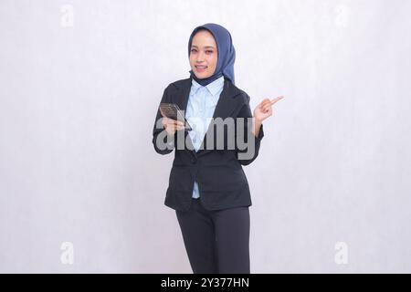 Jeune femme de bureau asiatique portant un hijab se tient heureux devant la caméra et pointe vers la gauche tout en portant un gadget de téléphone portable. Belle woma musulmane Banque D'Images