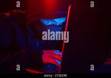 Cyber hacker dans le capot derrière l'ordinateur portable sur fond sombre avec la lumière bleue rouge, espion anonyme. concept de cybersécurité numérique Banque D'Images