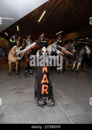 Un manifestant en costume tient un support de missile cassé lisant : « CESSEZ-LE-FEU MAINTENANT ! » Devant une ligne de chevaux de la police, Un blocus planifié et une manifestation contre les Forces terrestres 2024, la plus grande exposition de l'industrie de la défense dans l'hémisphère sud, ont éclaté en affrontements avec la police tout au long de la journée. Les manifestants qui tentaient d'empêcher l'entrée de la convention ont jeté des objets et des liquides sur les officiers, construit de petits barrages et déclenché des incendies d'ordures, tandis que la police a utilisé la force, le gaz poivre, les gaz lacrymogènes, les grenades paralysantes et les balles en caoutchouc sur les manifestants Banque D'Images