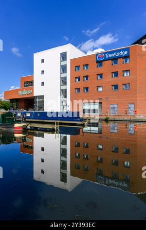 Grand Union canal dans le bassin d'Aylesbury, reflétant le supermarché Waitrose, l'hôtel Travelodge, des péniches et un bateau à moteur. Avec des eaux tranquilles et un ciel bleu. Banque D'Images