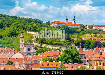 Le monastère de Strahov se trouve au sommet d'une colline surplombant la ville de Prague. Les toits orange et les tours jumelles se distinguent du feuillage vert luxuriant de la colline environnante. Ci-dessous, un réseau de toits de tuiles rouges s'étend vers l'horizon, formant une tapisserie vibrante et complexe. Le ciel au-dessus est une toile bleu vif, parsemée de nuages blancs moelleux. Banque D'Images