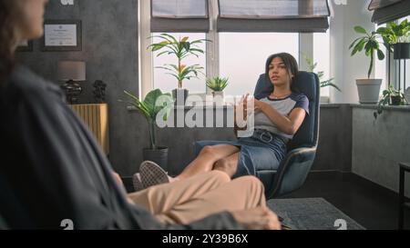 La jeune fille afro-américaine détendue est assise dans la chaise, partage des émotions positives avec le psychothérapeute, trouve la solution pendant la séance de thérapie de conseil. Psychothérapie et aide psychologique. La santé mentale. Banque D'Images