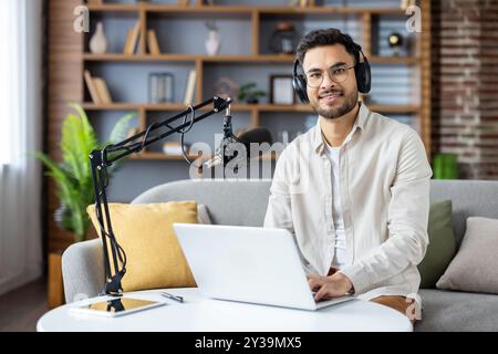 Podcaster souriant à l'aide d'un ordinateur portable et d'un microphone dans le home studio. Écouteurs, assis au bureau, créant du contenu audio avec enthousiasme. Une configuration moderne améliore l'expérience d'enregistrement, Banque D'Images