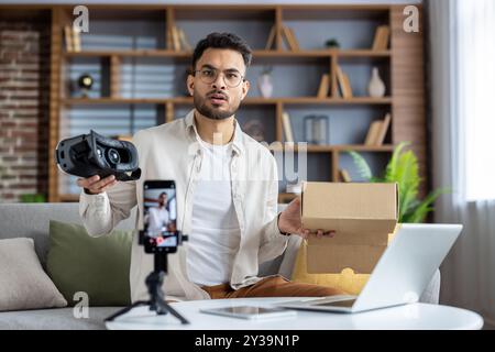 Portrait d'un jeune homme indien inquiet enregistrant un podcast et un blog au téléphone à la maison, assis sur un canapé et tenant une boîte à colis et un masque virtuel. Banque D'Images