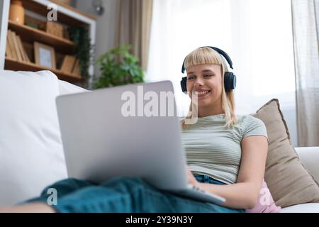 Jeune travailleur indépendant heureux ou femme étudiante à l'université assis travailler à la maison sur le canapé travaillant sur ordinateur portable, tout en écoutant podcast ou écouter la musique, Banque D'Images