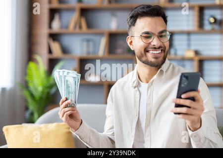 Homme souriant tenant des billets d'un dollar, utilisant un smartphone, exprimant le succès et la réussite financière. Dépeint le style de vie moderne, la technologie numérique, le gain financier. Banque D'Images